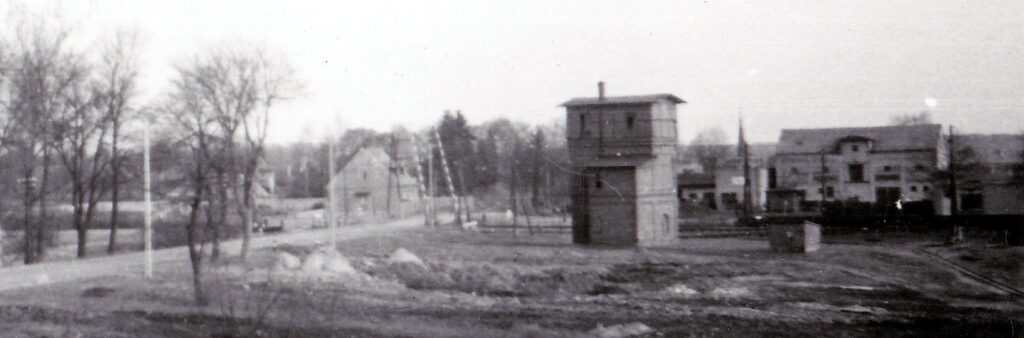 Widok trójkondygnacyjnej wieży ciśnień wzniesionej na planie prosotkąta, z przybudówką, na tle zabudowań przy ulicy Kolejowej w Człopie; po lewej stronie widoczne uniesione szlabany przejazdu kolejowego; drzewa w kadrze bez liści, ale i brak śniegu.