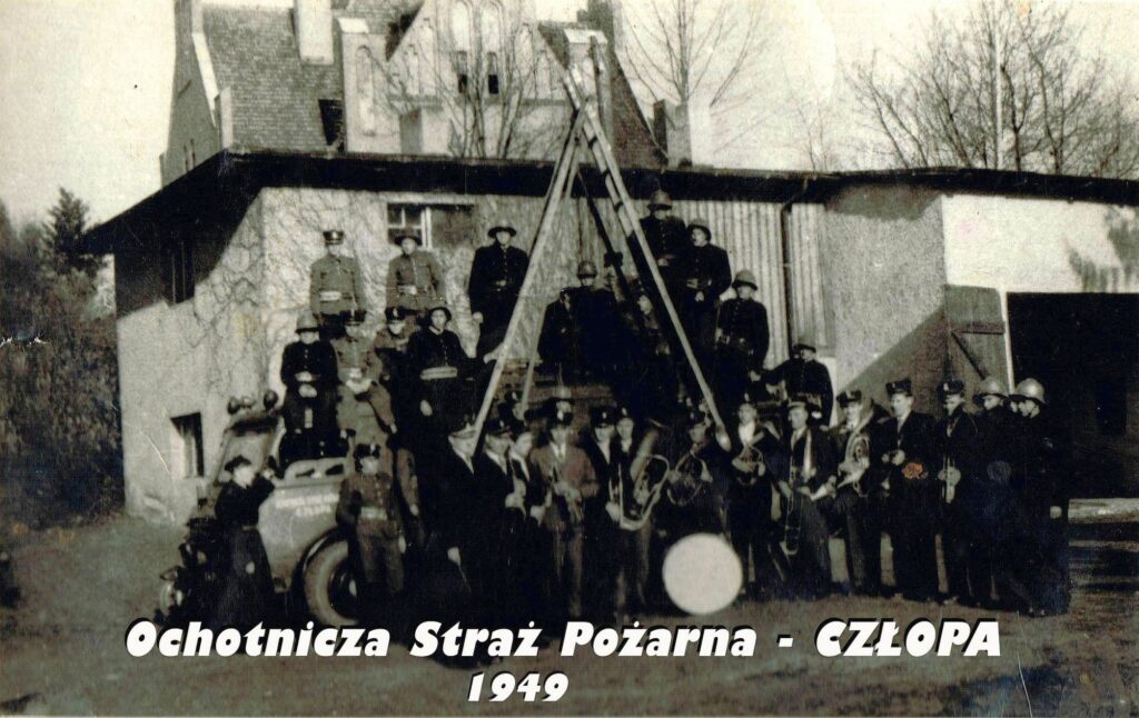 Fotografia zbiorowa członków jednostki OSP Człopa przed ich pierwszą remizą przy skrzyżowaniu ulic Stanisława Moniuszki i Strzeleckiej (w tle widoczny budynek dzisiejszego Urzędu Miasta i Gminy). Wóz gaśniczy „pozujący” wraz ze strażakami zaprzecza datacji zdjęcia na 1949 rok – to muszą już być co najmniej lata 1950. Warto zauważyć, że niektórzy obecni na zdjęciu trzymają instrumenty, co świadczy o istnieniu w tym czasie orkiestry strażackiej. Źródło fotografii: profil FB OSP Człopa, dostęp 3 maja 2026 roku – kliknij, aby przejść